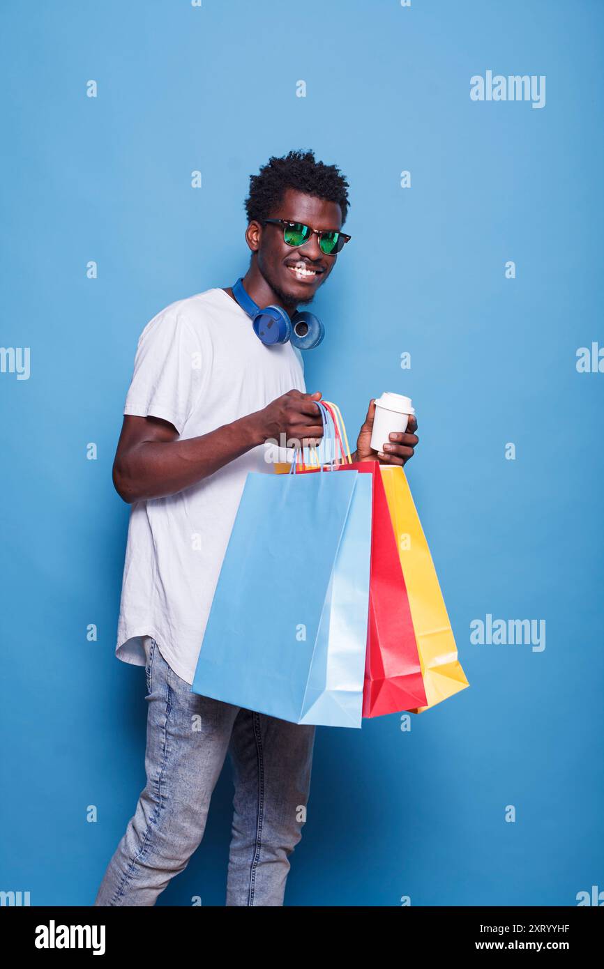Portrait d'homme noir portant des sacs à provisions et une tasse à café. Après avoir acheté des vêtements en vente, individu afro-américain insouciant portant des lunettes de soleil et souriant tout en tenant des sacs à vêtements. Banque D'Images