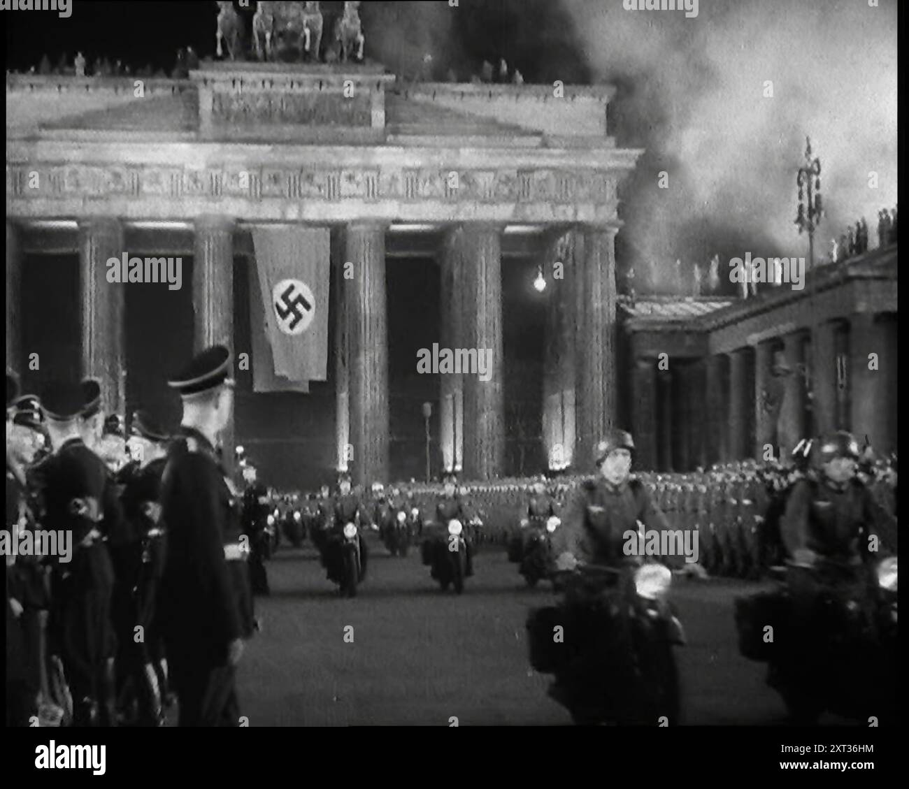 Officiers et soldats allemands qui bordent la Pariser Platz par la porte de Brandebourg avec un drapeau allemand accroché à elle comme une formation de Male Motorbike Riders Pass By, 1937. Berlin. 'Le leader [Adolf Hitler] revient de sa visite en Italie... on dit aux gens que le leader n'est pas seulement un homme, mais un Dieu sans faille, et qu'il doit être traité comme tel - et traité comme tel il est. Pas le retour d'un héros conquérant, car pour l'instant le héros n'a pas encore entrepris ses véritables conquêtes. Non, ce n'est que le retour du fils préféré de l'Allemagne. Quelque chose de nouveau. Quelque chose et quelqu'un d'une force motrice et d'influence. Ceci, à moins que Banque D'Images