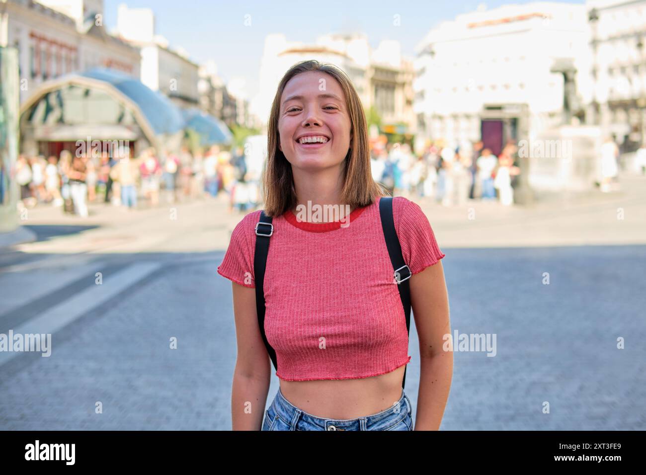 Une jeune femme, riant joyeusement, se tient sur une place animée de la ville regardant la caméra. Elle est habillée d'un short top rouge décontracté avec une sangle noire à l'arrière Banque D'Images