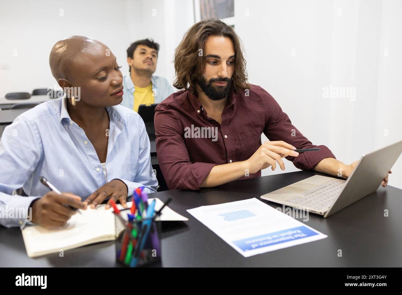 Dans une salle de classe moderne, un groupe diversifié de trois professionnels collabore sur un ordinateur portable pour discuter du travail académique, mettant en évidence le travail d'équipe dans un Banque D'Images