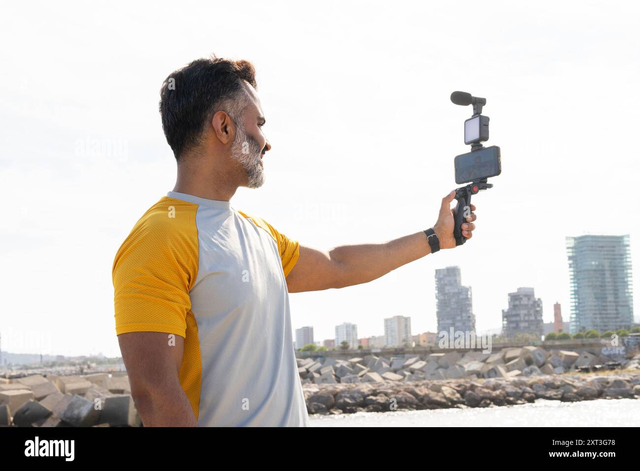 Homme en t-shirt jaune utilisant une nacelle avec smartphone pour enregistrer une vidéo au bord de la mer, regardant l'écran, avec skyline urbaine en arrière-plan. Banque D'Images