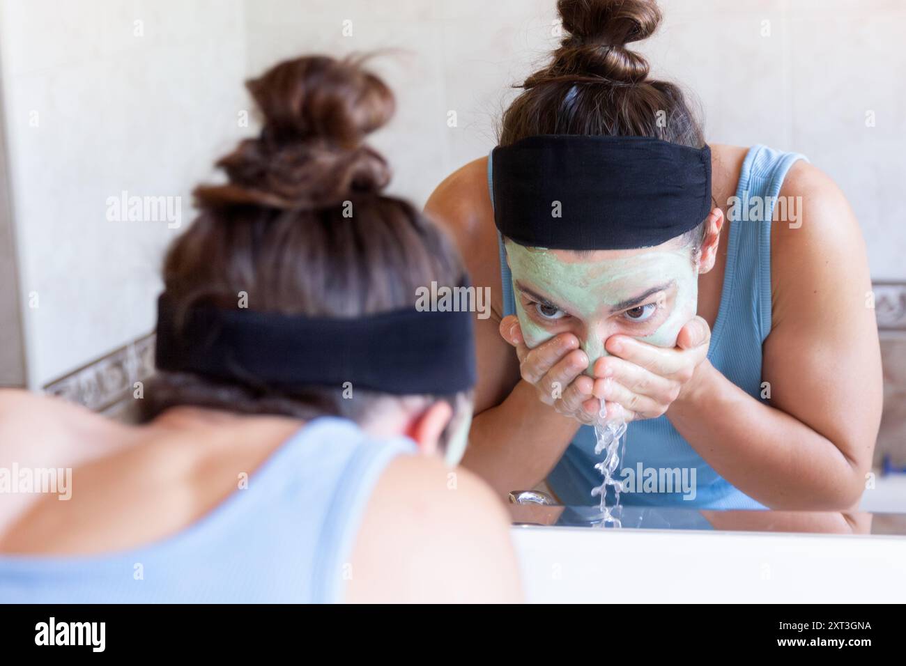 Une jeune femme avec un bandeau lave soigneusement un masque vert sur son visage dans une salle de bain bien éclairée, en se concentrant sur la santé de sa peau Banque D'Images