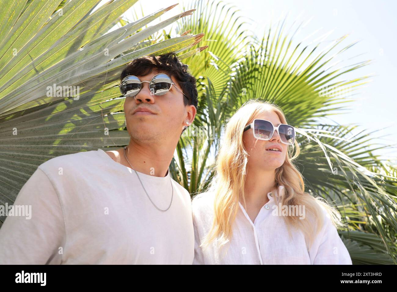 Un jeune couple élégant portant des lunettes de soleil se tient sous les frondes de palmier, se prélasser dans la lumière du soleil, exsudant l'élégance décontractée et les vibrations estivales Banque D'Images