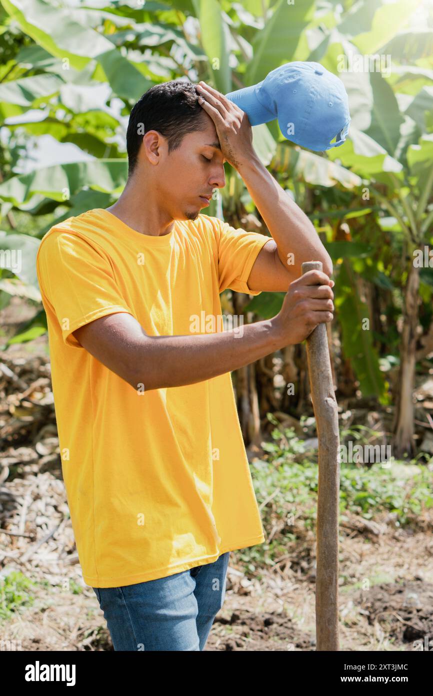 Un jeune homme vêtu d'un t-shirt jaune vif et d'un Jean se tient debout dans un jardin ensoleillé, essuyant la sueur de son front et tenant un outil de jardinage Banque D'Images