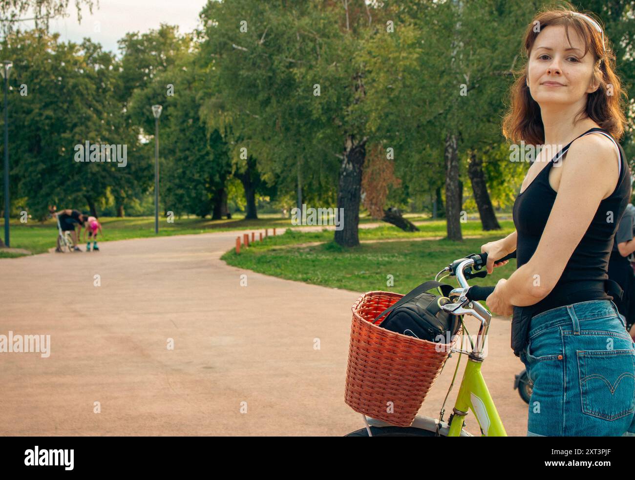 Jeune femme avec vélo dans l'allée. Belle femme en vêtements décontractés debout avec vélo avec panier. Femme active dans un parc public. Banque D'Images