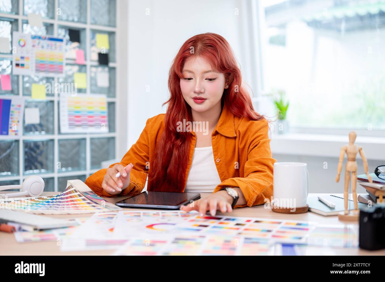 Une graphiste féminine asiatique belle et créative avec les cheveux bouclés rouges travaille à son bureau dans le studio, sélectionnant des couleurs à partir d'un ensemble de couleurs Banque D'Images