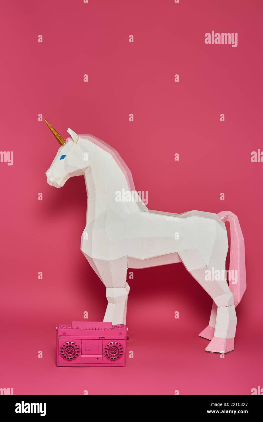 Une licorne en origami blanc se tient devant un fond rose avec une boombox rose à ses pieds. Banque D'Images