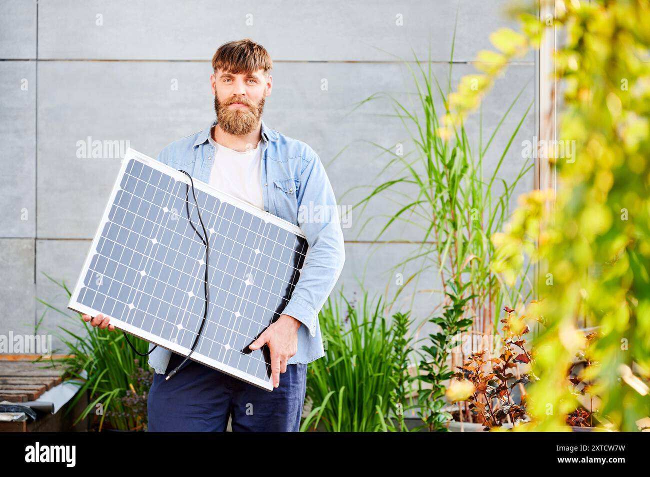 Portrait de l'homme barbu tient un panneau solaire, debout dans un cadre moderne avec des plantes et de la verdure. Guy en chemise bleu clair et t-shirt blanc semble heureux et détendu. Banque D'Images