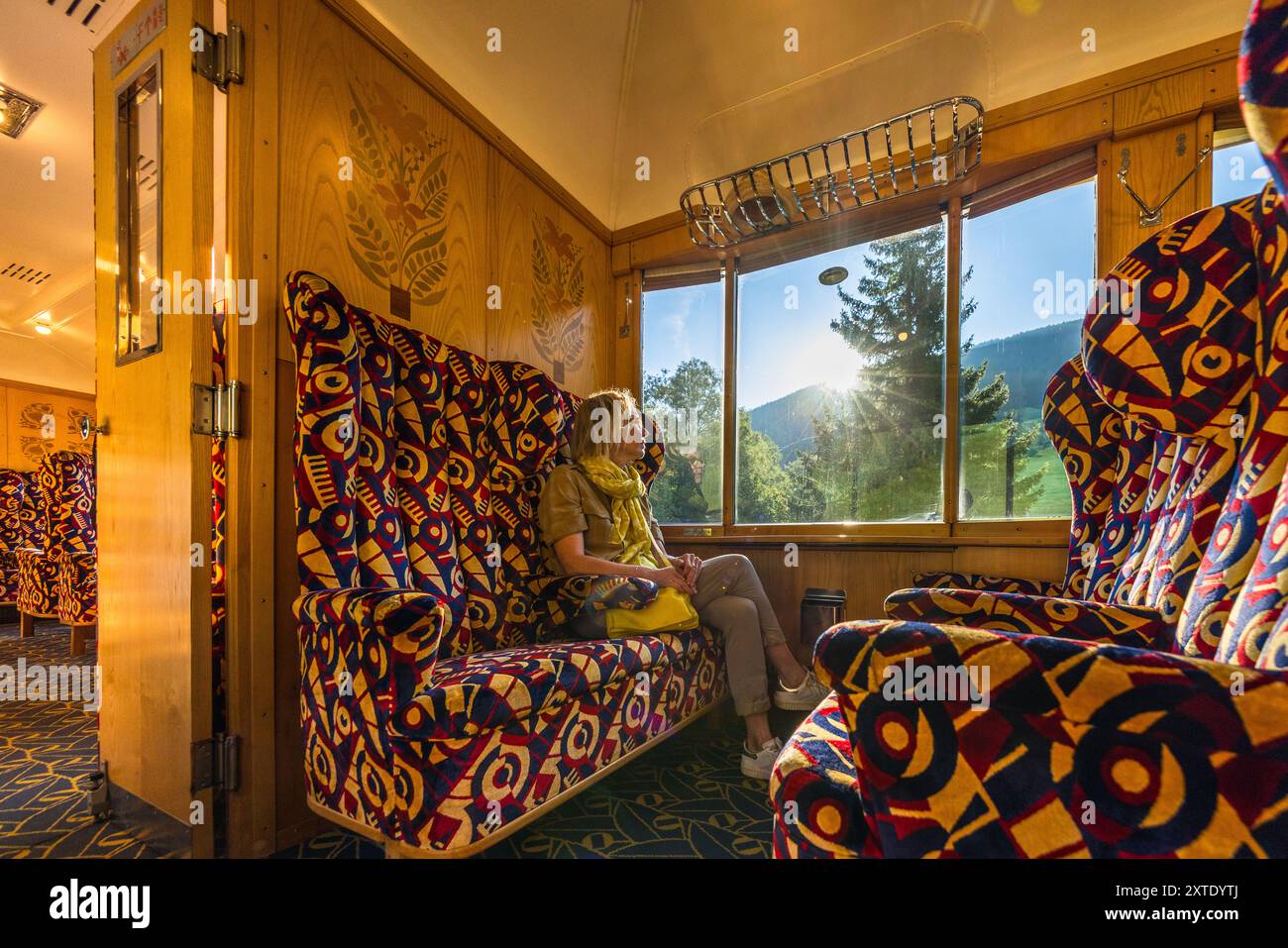 La journaliste Angela Berg est assise près de la fenêtre d’un wagon du Alpine Classic Pullman Express, contemplant le paysage qui défile. Le train date de 1931 et circulait à l’origine sous le nom de « Golden Mountain Pullman Express » sur la ligne ferroviaire Montreux–Berner Oberland. Le chemin de fer rhétique l’a acquis en 1939, et le designer français René Prou avait créé les incrustations en bois et les revêtements Art Déco des wagons. Le Verein Pro Salonwagen, fondé en 1998, a sauvé le train, qui est aujourd’hui un monument ferroviaire historique protégé dans le canton des Grisons. Talstrasse, Davos, Grisons, Suisse. Banque D'Images