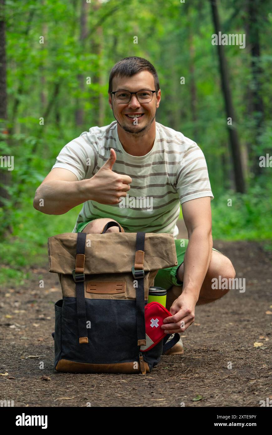 Randonneur souriant donnant un pouce vers le haut tout en tenant une trousse de premiers soins à côté de son sac à dos sur un sentier forestier, montrant la préparation et l'expérience positive en plein air. Banque D'Images