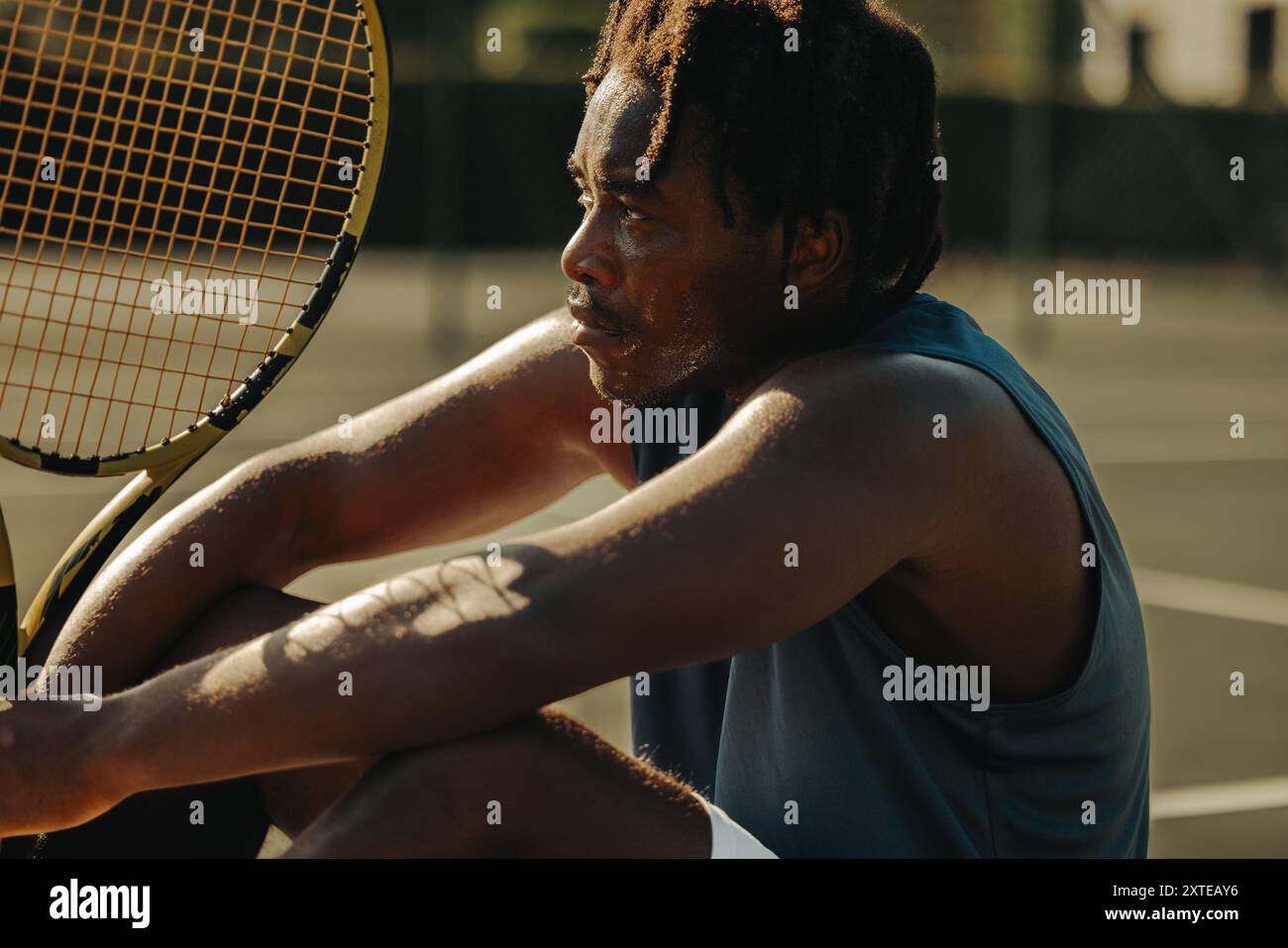 Homme afro-américain avec des dreadlocks assis sur un court de tennis tenant une raquette, capturant un moment de repos pendant un match. Banque D'Images