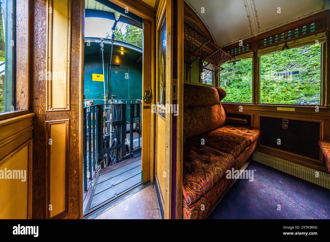 La vue depuis un compartiment lambré d’un wagon ferroviaire historique passe par la porte de communication ouverte sur la plateforme d’attelage du wagon suivant, vêtu de vert, entouré d’une forêt dense, ligne de l’Albula, Grisons. Le Chemin de fer rhétique assure ce trajet aller-retour Davos Platz–Filisur–Davos Platz avec la locomotive Ge 4/4 I n° 605 « Silvretta », mise en service en 1953. La ligne de l’Albula fait partie du site du patrimoine mondial de l’UNESCO « Chemin de fer rhétique dans les paysages de l’Albula/Bernina » depuis 2008, Landwasserstrasse, Davos, Suisse. Banque D'Images