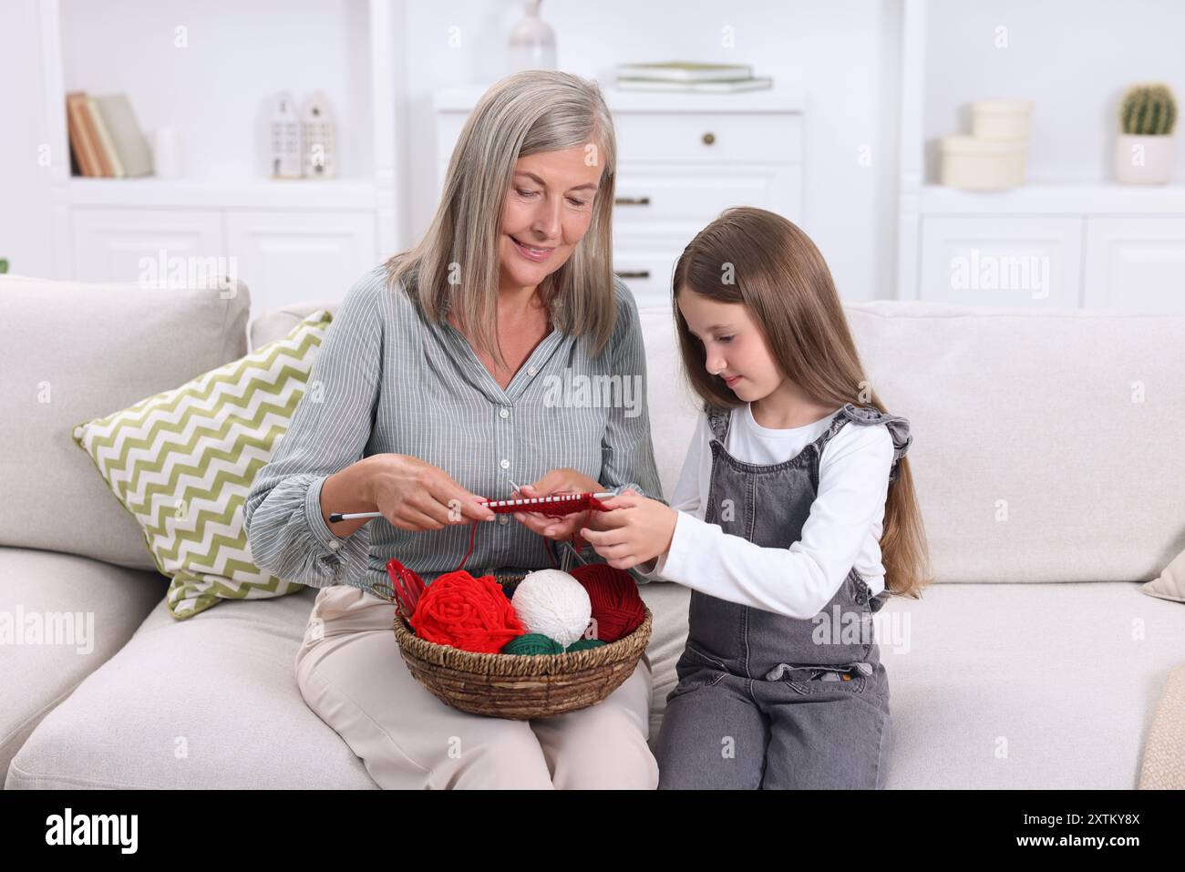 Grand-mère souriante apprenant à sa petite-fille à tricoter sur le canapé à la maison Banque D'Images