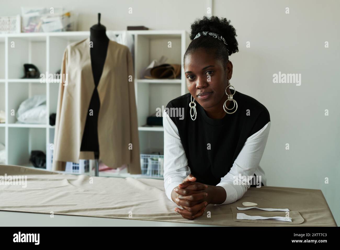 Jeune tailleur afro-américaine ou créatrice de mode regardant la caméra tout en se tenant debout près de la table avec du textile gris déroulé Banque D'Images