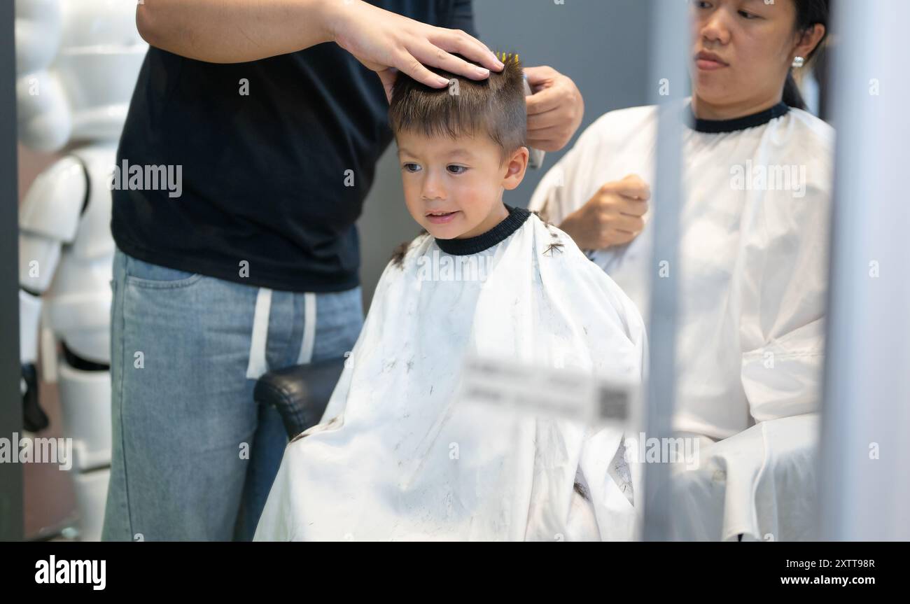 Un jeune garçon sourit alors qu'il se fait couper les cheveux par un coiffeur doux dans un salon contemporain. La scène capte un moment de bonheur et de confiance, de réflexion Banque D'Images