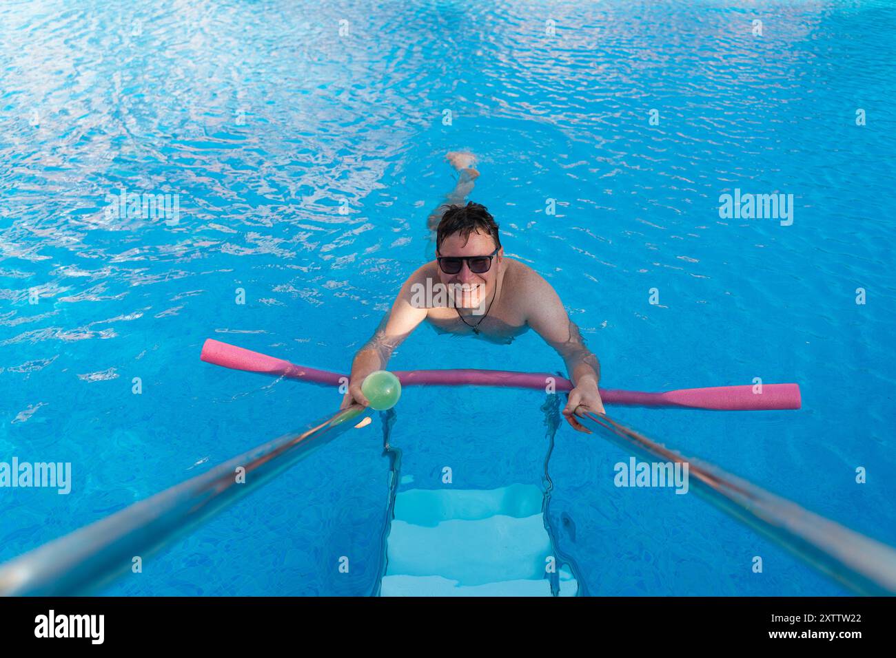 Un homme profite d'une journée ensoleillée au bord de la piscine, jouant avec un jouet de nouilles roses. Il est vu souriant, nageant et se relaxant dans l'eau bleue claire. Extérieur Banque D'Images