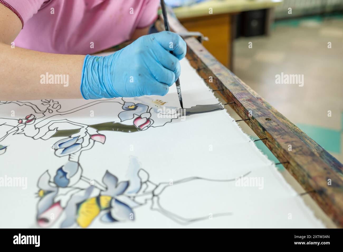 Dans un studio serein, une artiste féminine qualifiée dans un gant bleu peint méticuleusement des fleurs blanches complexes sur un tissu de soie bleue luxueux, créant un capt Banque D'Images
