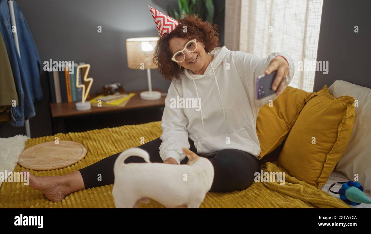 Une femme mature prend un selfie sur son lit avec son chihuahua animal de compagnie dans une chambre confortable, portant un chapeau de fête et des lunettes. Banque D'Images