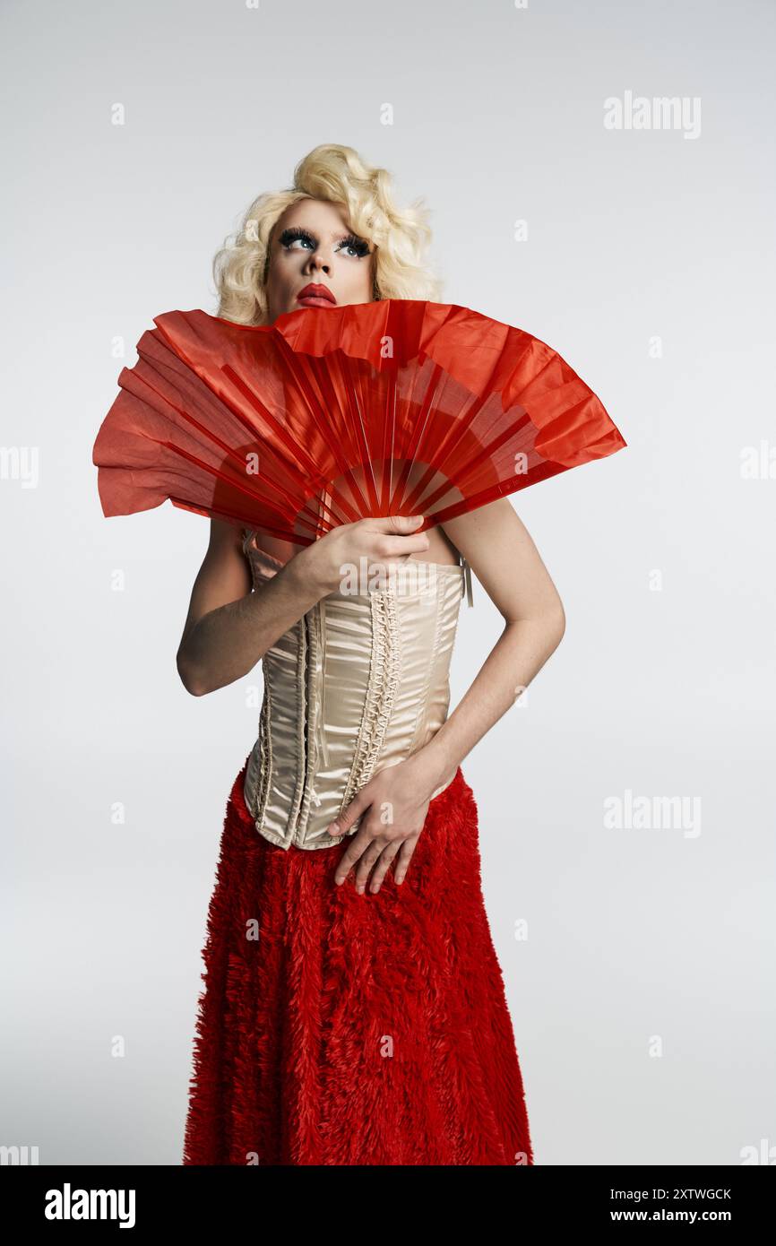 Une drag queen dans une perruque blonde, une jupe rouge et un corset pose dramatiquement, tenant un éventail rouge devant elle. Banque D'Images