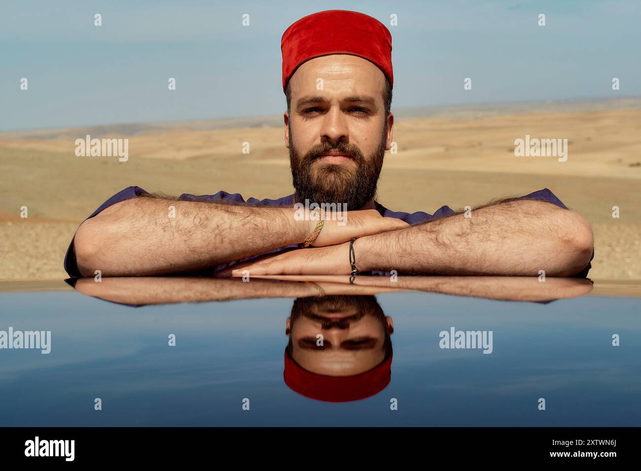 Homme à la barbe portant un chapeau rouge de fès repose son menton sur ses bras croisés, reflété sur une surface brillante sur un fond désert. Banque D'Images