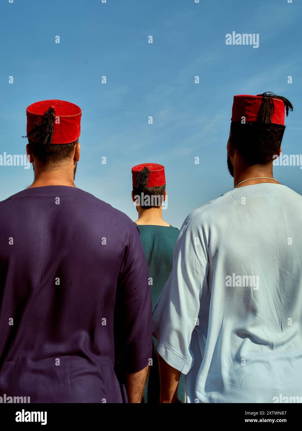 Trois individus portant des chapeaux rouges traditionnels et des vêtements colorés debout contre un ciel bleu clair. Banque D'Images