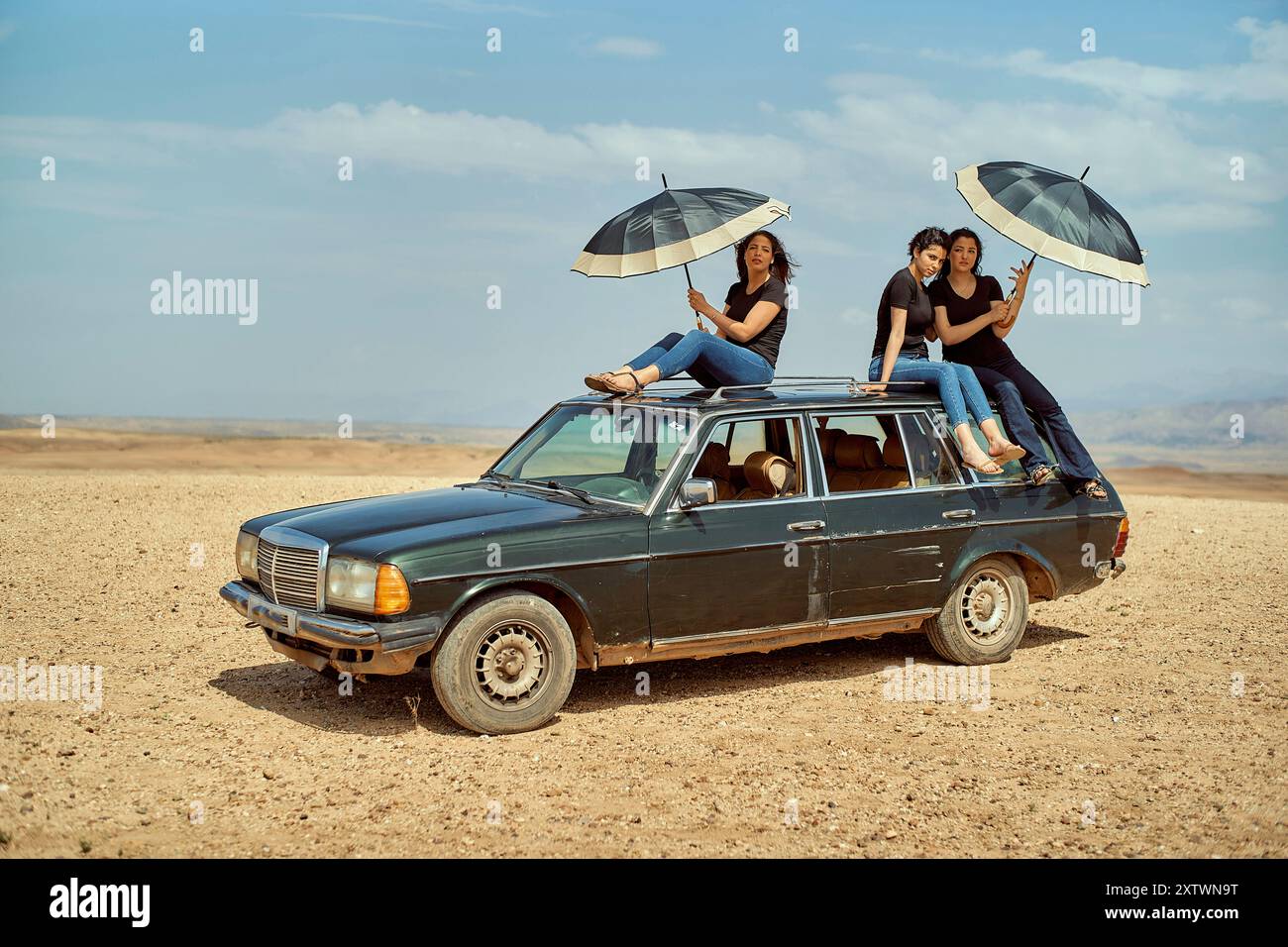 Groupe d'amis s'amusant avec des parapluies sur une voiture vintage dans un cadre désertique. Banque D'Images