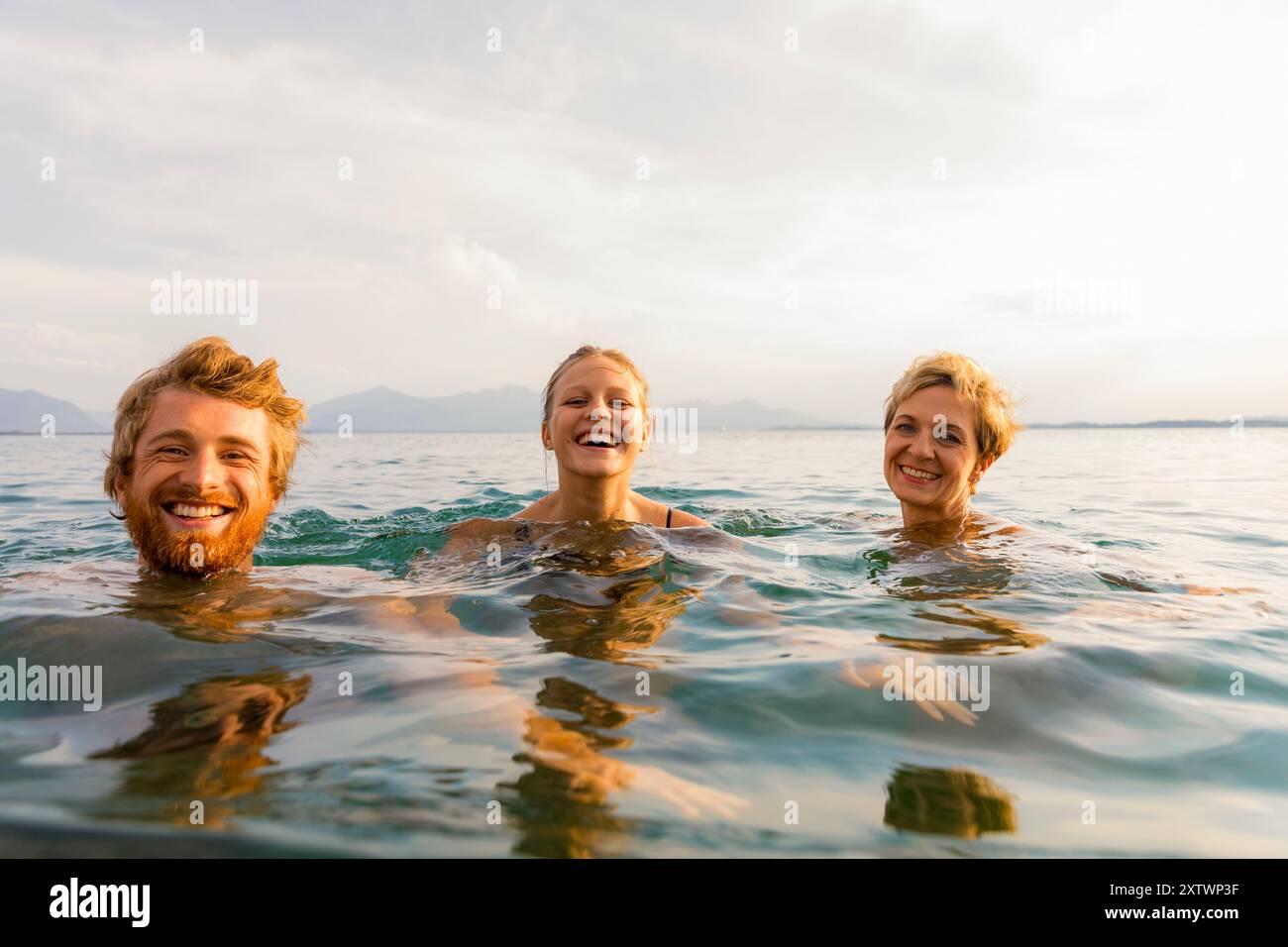 Trois personnes sourient et nagent dans un cadre d'eau libre avec des montagnes en arrière-plan sous un ciel clair. Banque D'Images