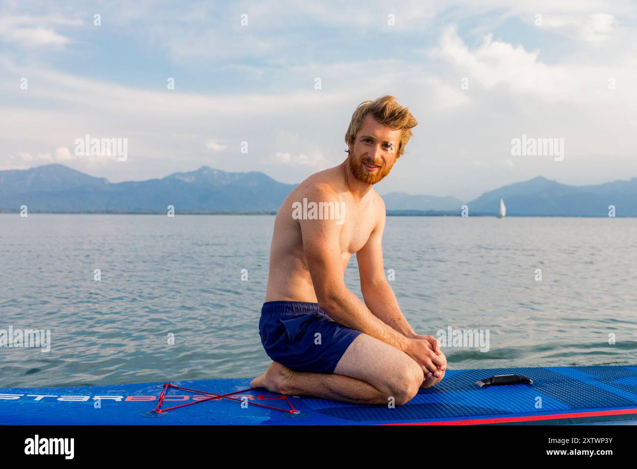 Homme souriant avec une barbe assise sur une planche à paddleboard sur un lac calme avec des montagnes en arrière-plan. Banque D'Images