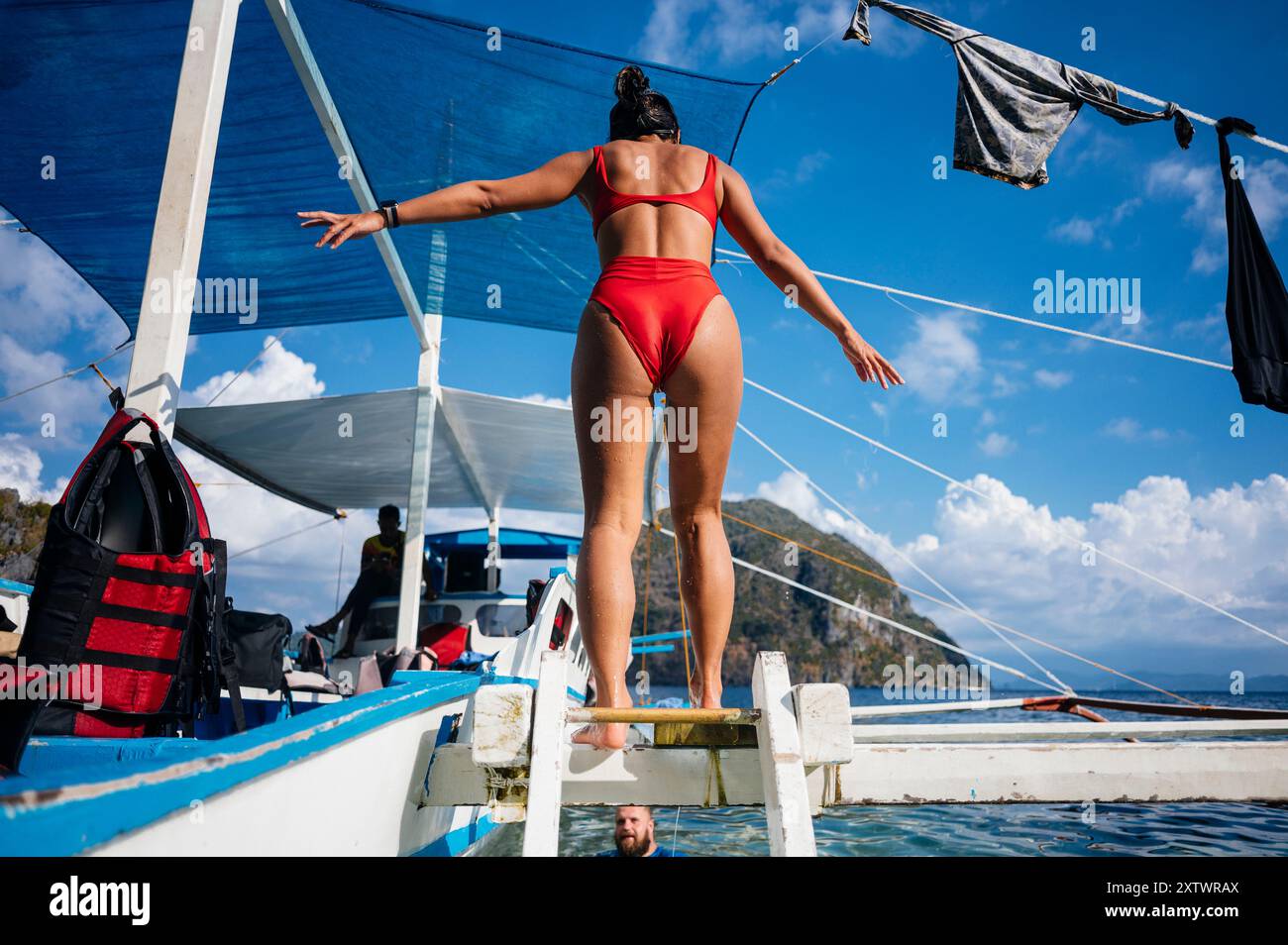 Femme dans un bikini rouge sur le point de plonger dans la mer d'un bateau avec une bâche bleue et un ciel clair en arrière-plan. Banque D'Images
