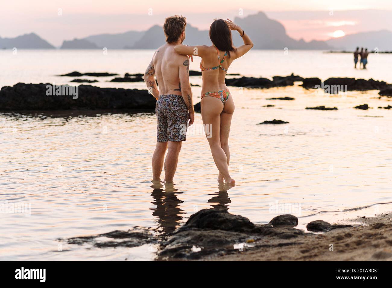 Un couple se tient la main tout en marchant sur une plage au coucher du soleil, avec l'océan lançant doucement à leurs pieds et les montagnes au loin. Banque D'Images