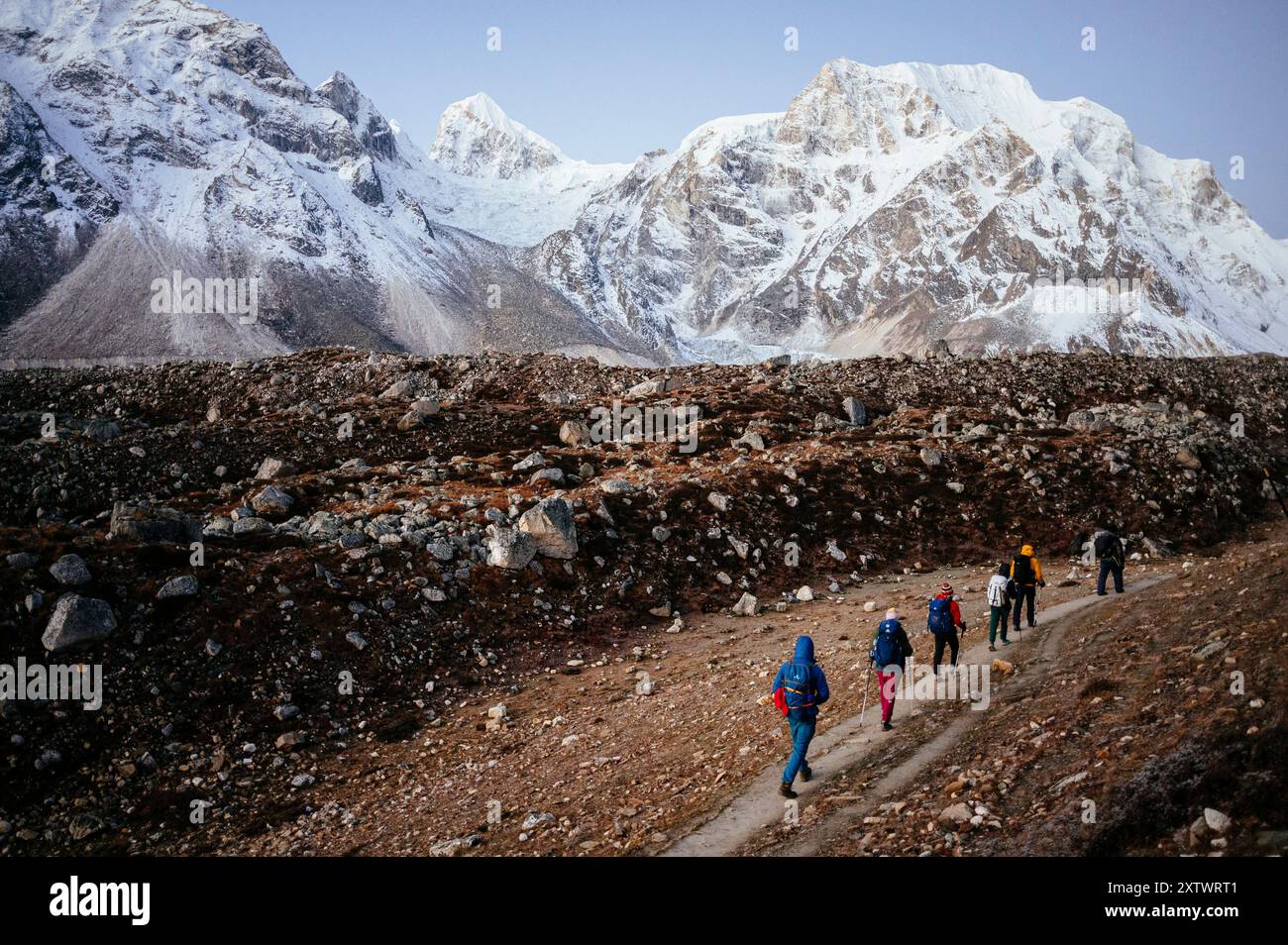 Un groupe de randonneurs marchent le long d'un sentier de montagne avec des sommets enneigés en arrière-plan sous un ciel matinal clair. Banque D'Images