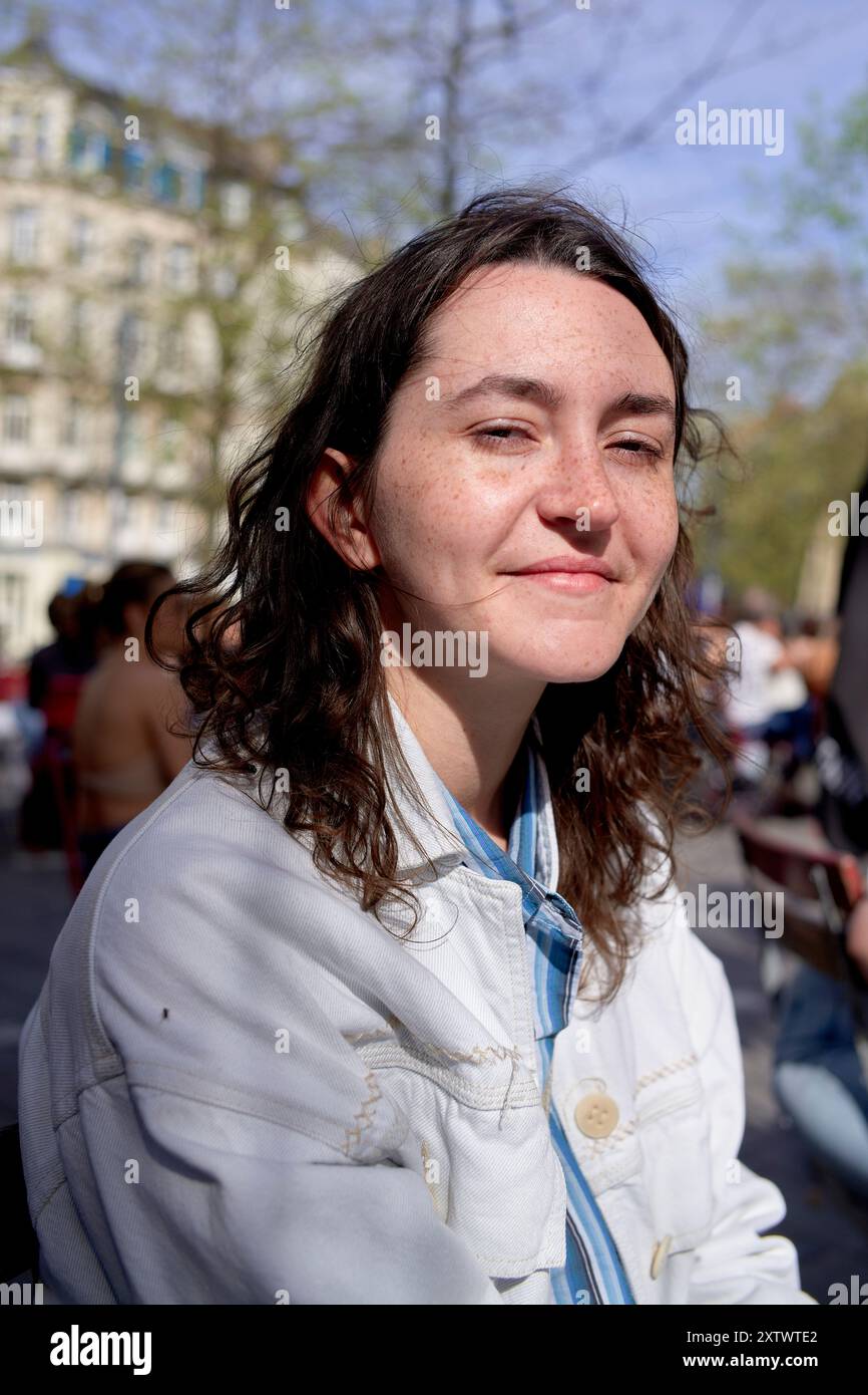 Femme souriante avec les cheveux bruns assis à l'extérieur dans une veste blanche par une journée ensoleillée. Banque D'Images