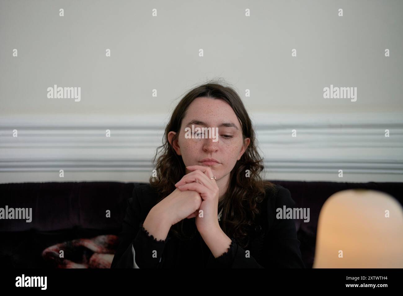 Femme assise sur un canapé profondément dans la pensée avec ses mains serrées, une lampe floue au premier plan. Banque D'Images