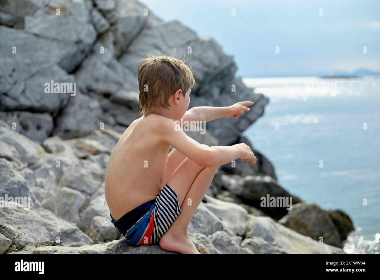 Un jeune garçon en maillot de bain rayé est assis sur des rochers près de la mer, pointant vers l'horizon. Banque D'Images