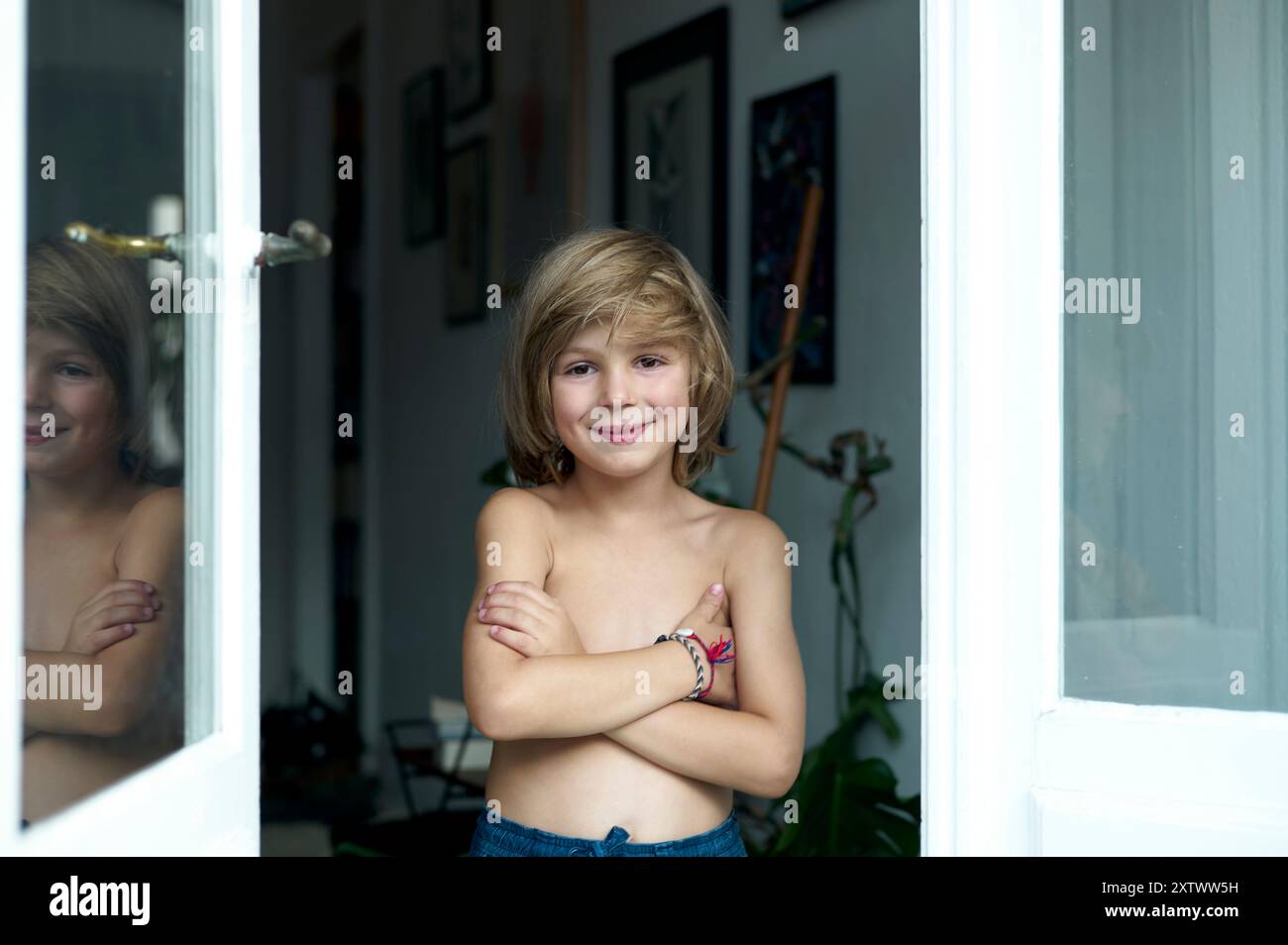 Enfant souriant avec des cheveux blonds debout par une fenêtre avec les bras croisés, reflétant dans la vitre. Banque D'Images