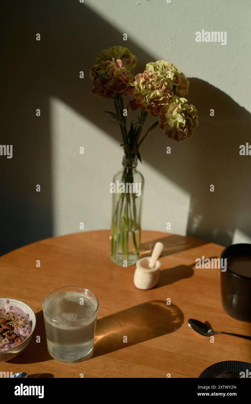 Table en bois ensoleillée avec un vase de fleurs, un verre d'eau et un bol avec des ustensiles. Banque D'Images