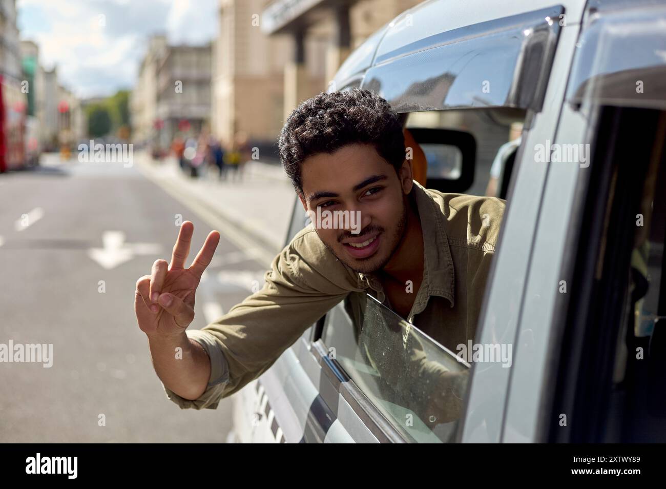 Jeune homme souriant donnant un signe de paix du côté conducteur d'une voiture argentée dans une rue ensoleillée de la ville. Banque D'Images