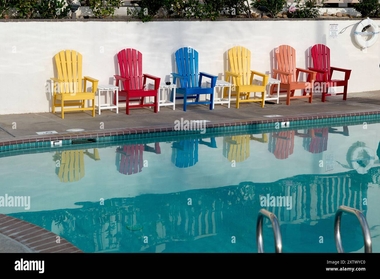 Chaises Adirondack colorées alignées à côté d'une piscine calme reflétant les chaises et le ciel bleu clair. Banque D'Images