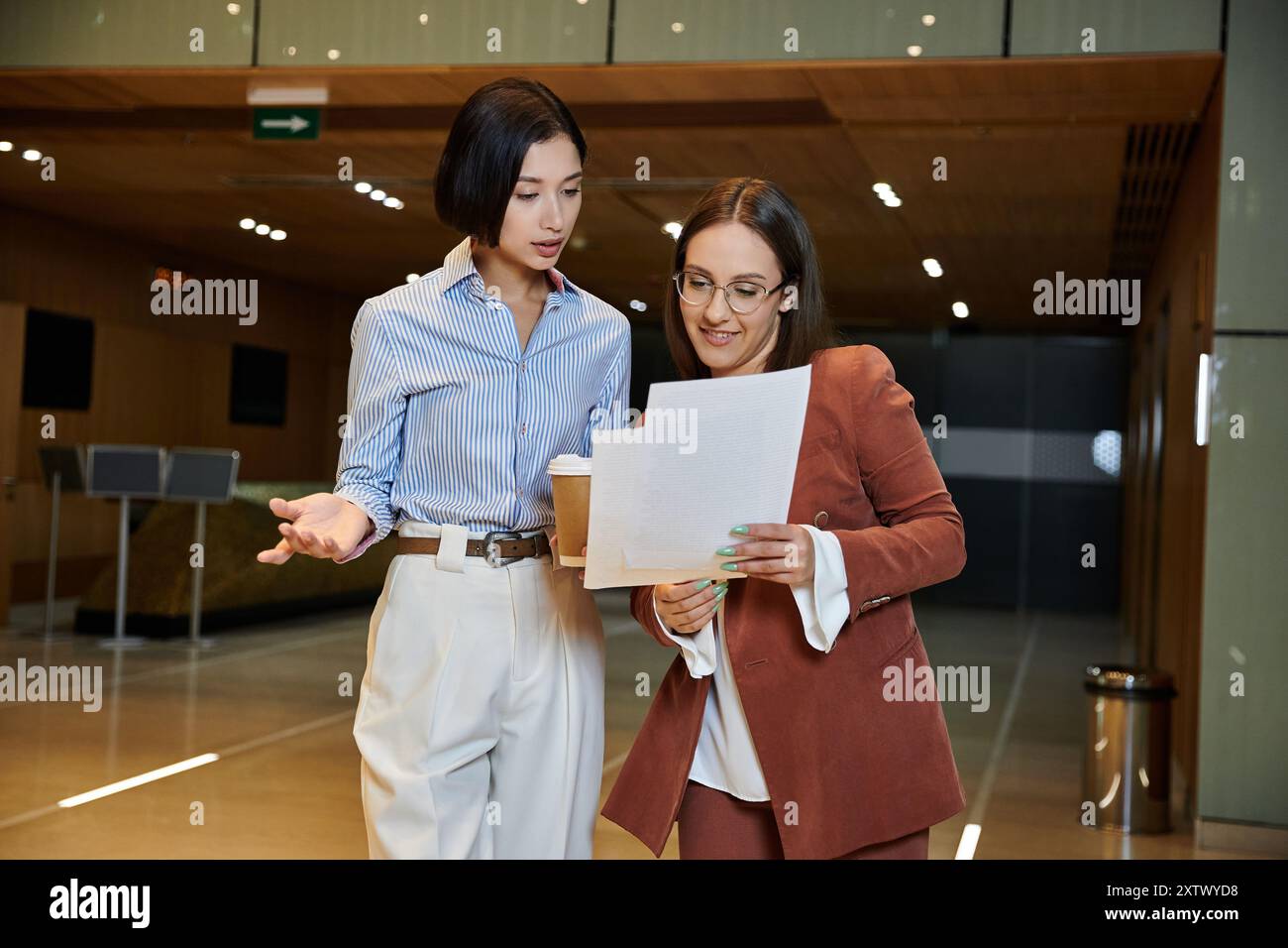 Deux femmes en tenue professionnelle conversent et examinent des documents dans un hall de bureau moderne. Banque D'Images