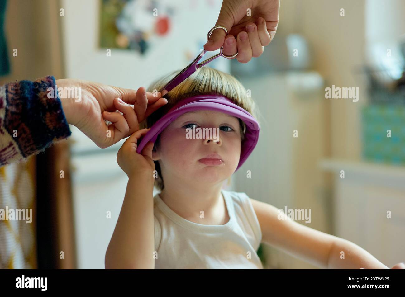 Un enfant s'assoit patiemment tandis qu'une main adulte coupe ses cheveux avec des ciseaux, un bandeau violet est utilisé pour garder les cheveux en place. Banque D'Images