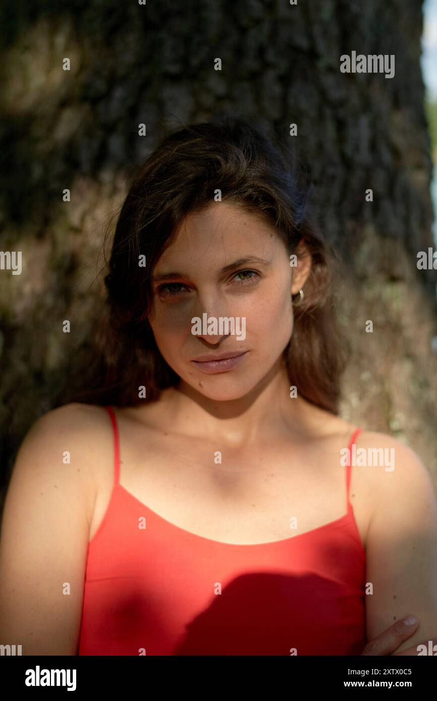 Une femme confiante dans une robe rouge posant devant un arbre avec la lumière du soleil tapissée illuminant son visage. Banque D'Images