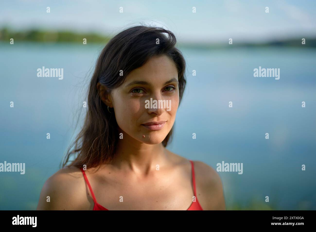 Une femme au sommet rouge se tient au bord d'un lac avec une expression sereine sur son visage. Banque D'Images