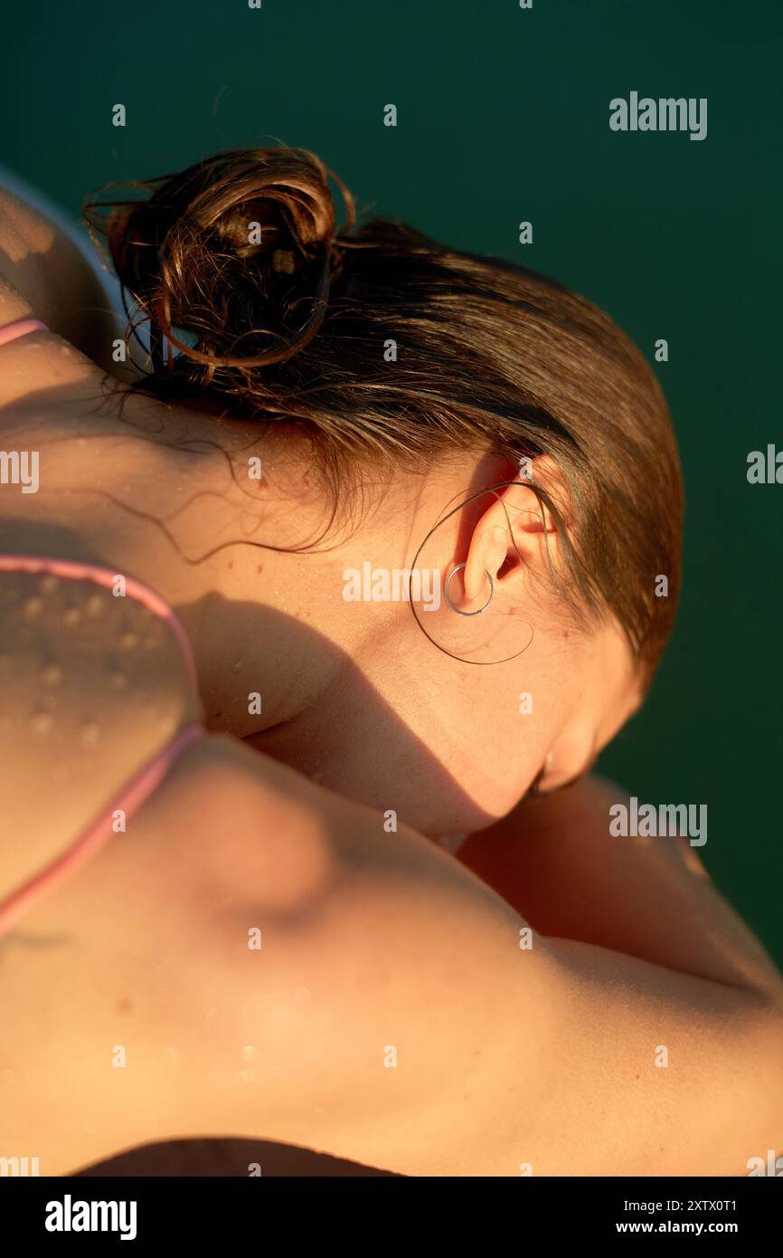 Profil embrassé par le soleil d'une personne avec les cheveux attachés, montrant une gouttelette d'eau sur l'épaule sur un fond sarcelle. Banque D'Images