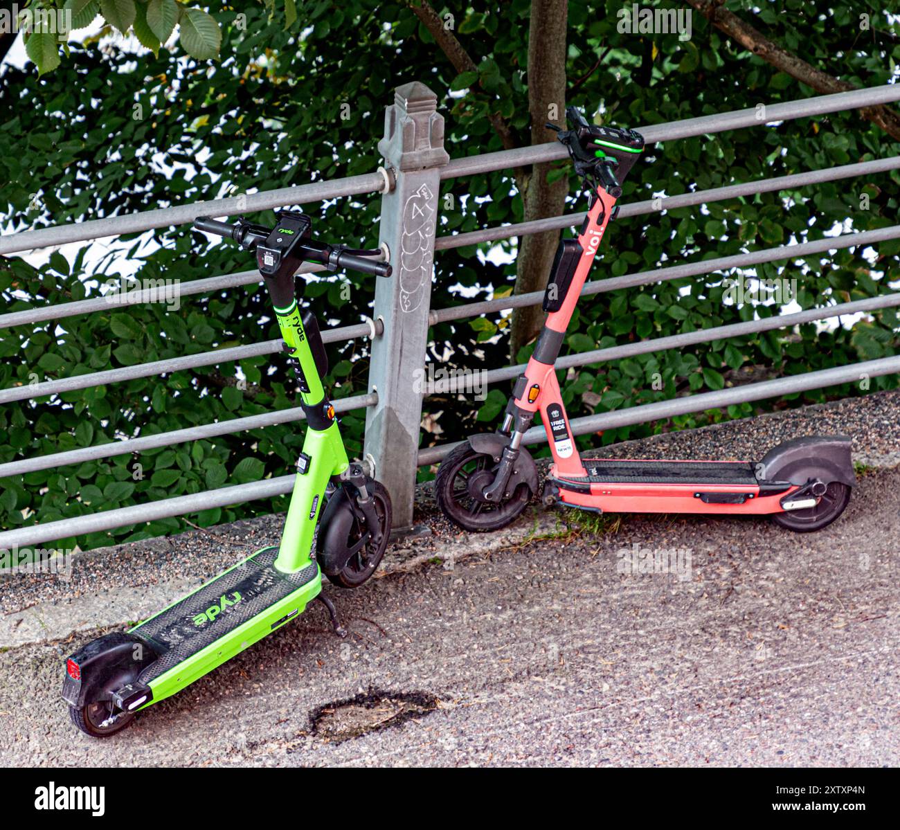 Un Ryde et un scooter électrique Voi garés dans une rue, face à face, à côté d'une clôture. Arbres en arrière-plan. Banque D'Images