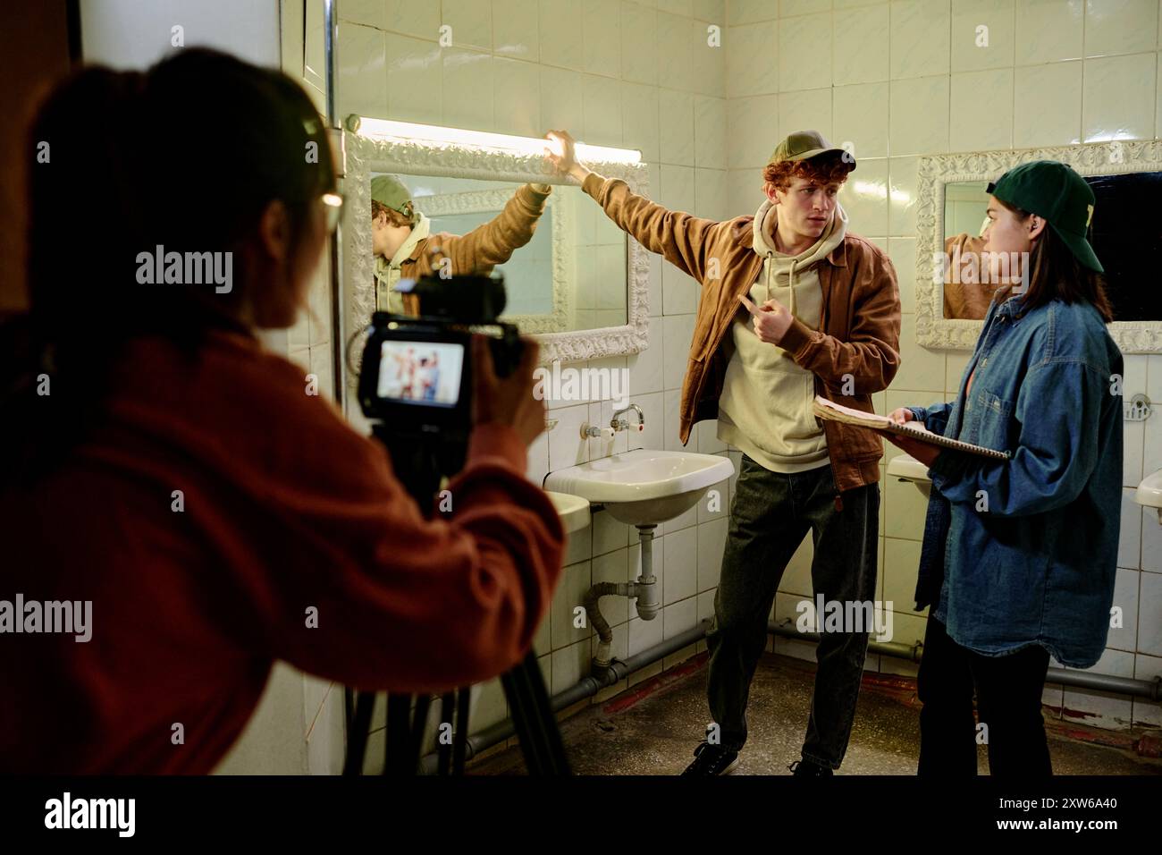 Deux acteurs debout dans une salle de bains carrelée avec lavabos, miroirs et équipement de tournage tout en jouant la scène. Cameraperson en arrière-plan capturant le moment Banque D'Images