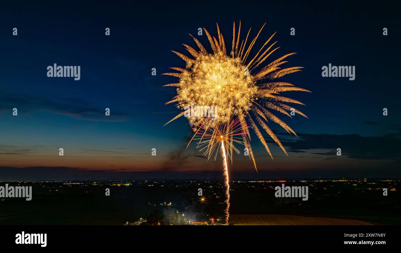Des feux d'artifice dorés explosent dans le ciel nocturne, formant Un grand éclat radieux au-dessus des lumières lointaines de la ville et Une foule rassemblée, avec l'horizon crépusculaire rehaussant l'atmosphère festive. Banque D'Images