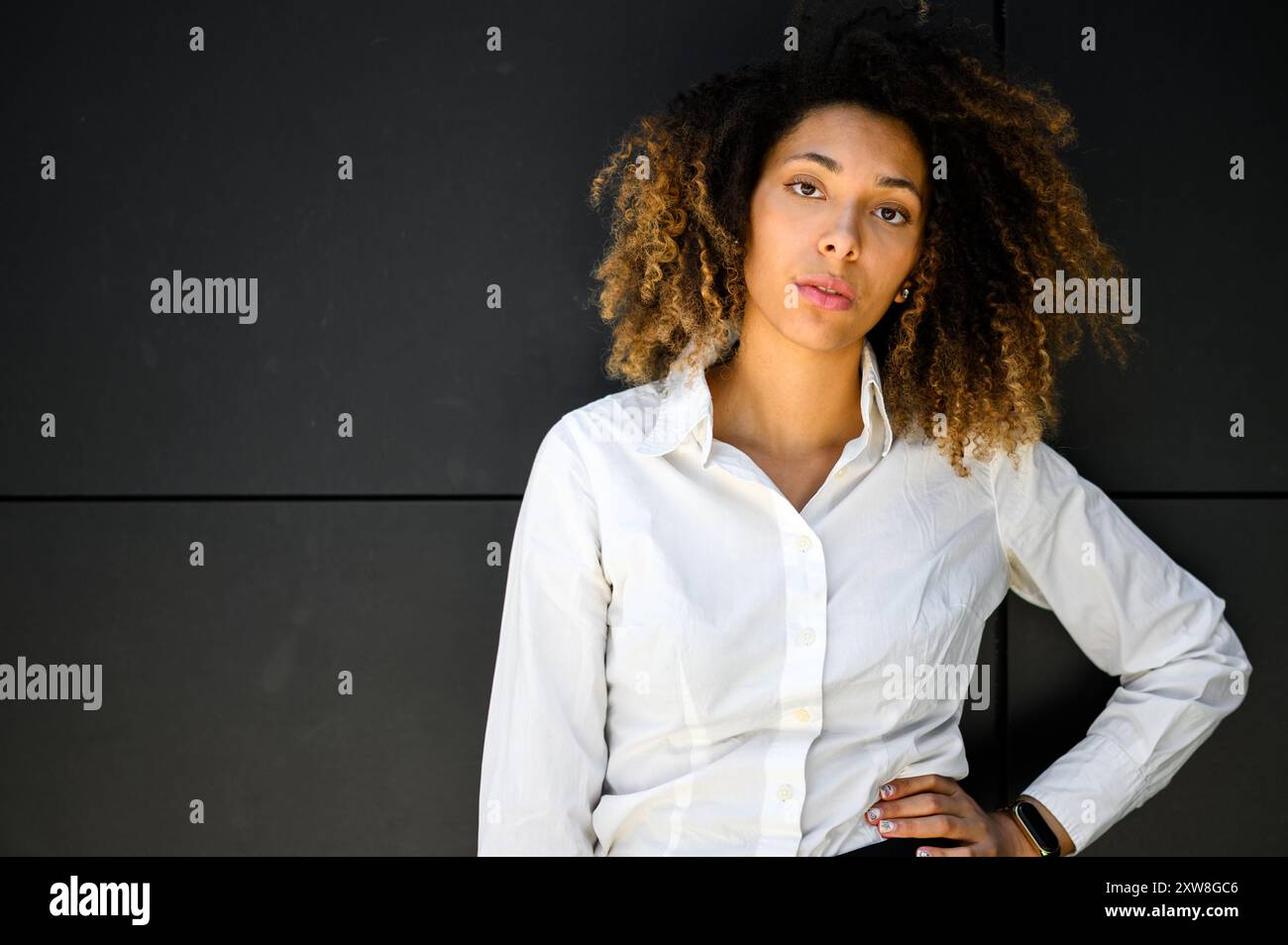 Une jeune femme professionnelle aux cheveux bouclés se tient devant un mur noir moderne, porte une chemise blanche et pose avec sa main sur sa hanche Banque D'Images