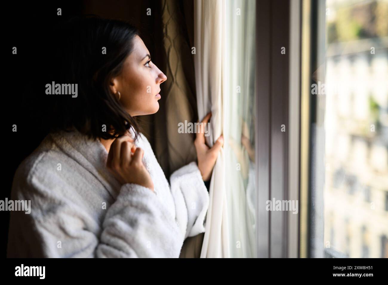 Jeune femme pensive dans une robe blanche confortable regarde par une fenêtre, profondément dans la pensée Banque D'Images