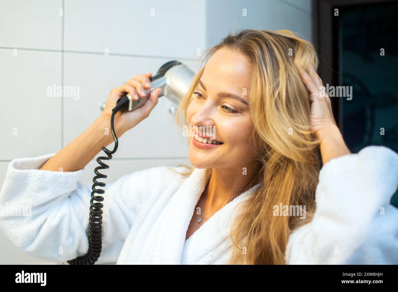 Jeune femme joyeuse dans une robe blanche coiffe ses cheveux blonds avec un sèche-cheveux dans une salle de bain lumineuse, incarnant self-Care et routine de beauté quotidienne Banque D'Images