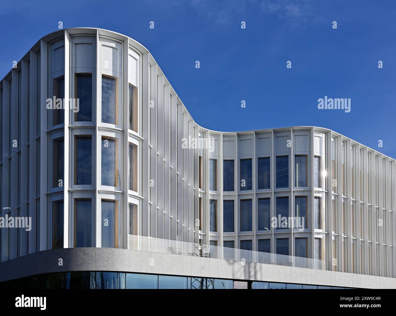 L'image met en valeur un bâtiment architectural moderne avec une façade élégante et ondulée. La structure comporte de grandes fenêtres alignées verticalement encadrées par un Banque D'Images